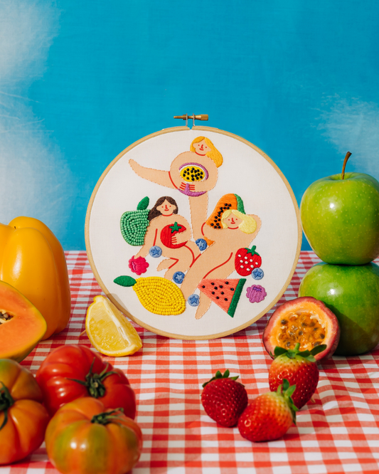 Embroidery hoop with colorful fruit-themed design on a table with fruits and vegetables.