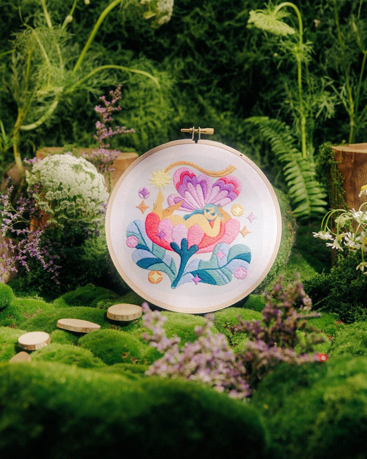 A round embroidery hoop displaying a colorful floral design, set in a lush, mossy garden scene with wildflowers and green foliage.