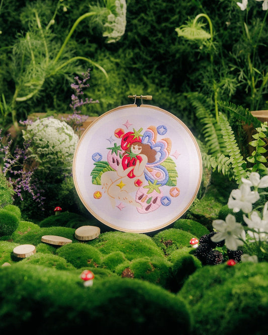 A round embroidery hoop displaying a colorful floral and whimsical design, set against a lush, miniature garden backdrop of green moss, ferns, white flowers, and tiny stones.