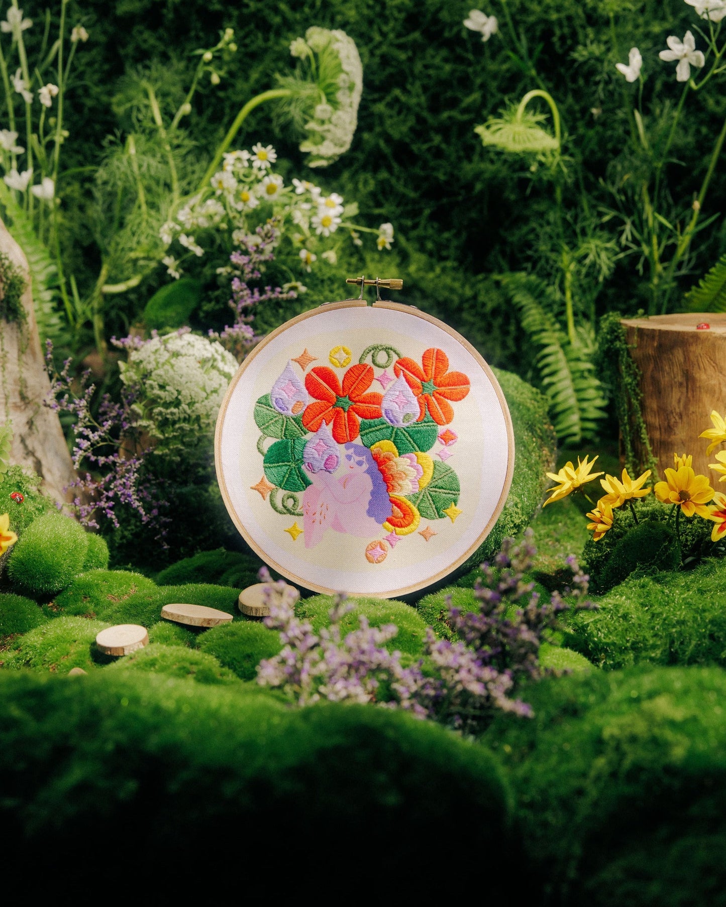 A round decorative plate with a colorful floral and butterfly illustration, displayed upright in a lush green garden setting with moss, flowers, and a small wooden planter.