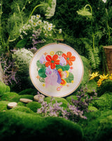 A round decorative plate with a colorful floral and butterfly illustration, displayed upright in a lush green garden setting with moss, flowers, and a small wooden planter.