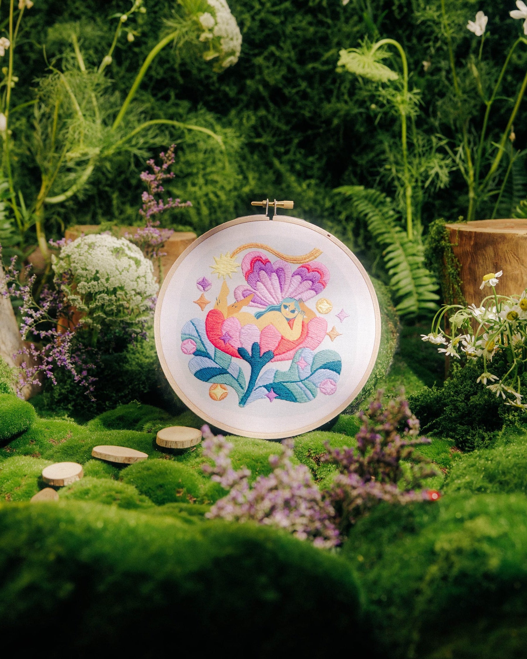 A round embroidery hoop displaying a colorful floral design, set in a lush, mossy garden scene with wildflowers and green foliage.