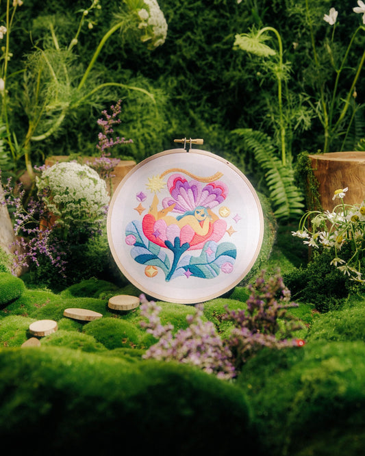 A round embroidery hoop displaying a colorful floral design, set in a lush, mossy garden scene with wildflowers and green foliage.