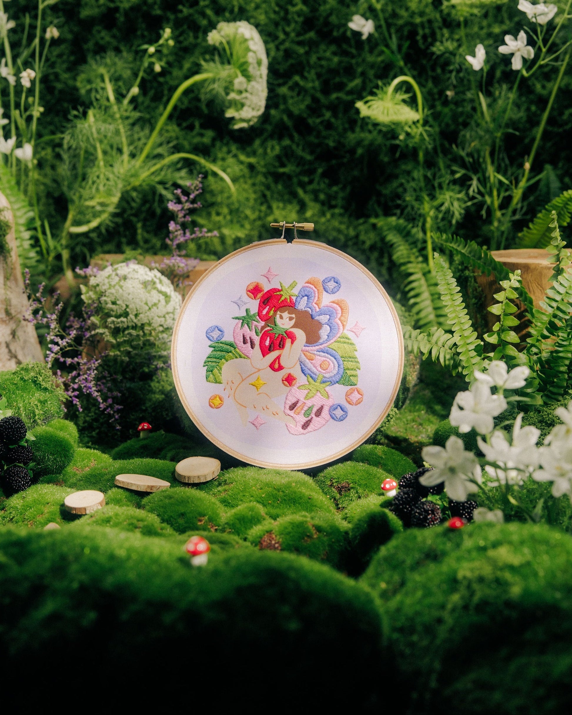 A round embroidery hoop displaying a colorful floral and whimsical design, set against a lush, miniature garden backdrop of green moss, ferns, white flowers, and tiny stones.